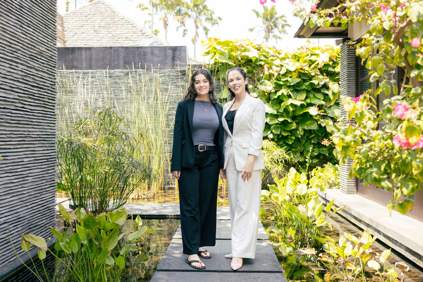 Two professionally dressed women stand together on a stone walkway in a lush outdoor garden with a water feature behind them.